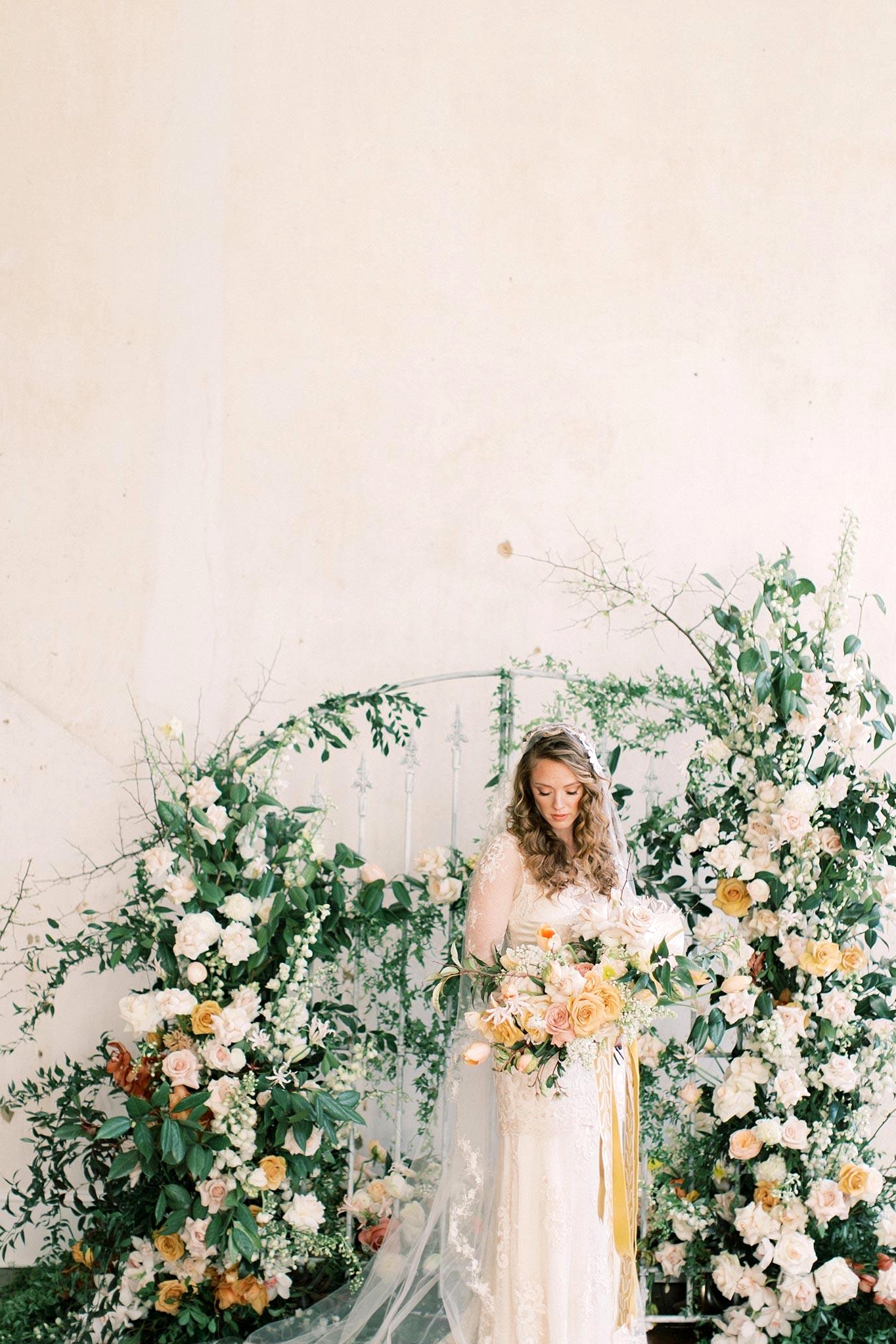 lush floral gate wedding backdrop with bride in a vintage-inspired wedding dress holding a yellow and white spring bridal bouquet