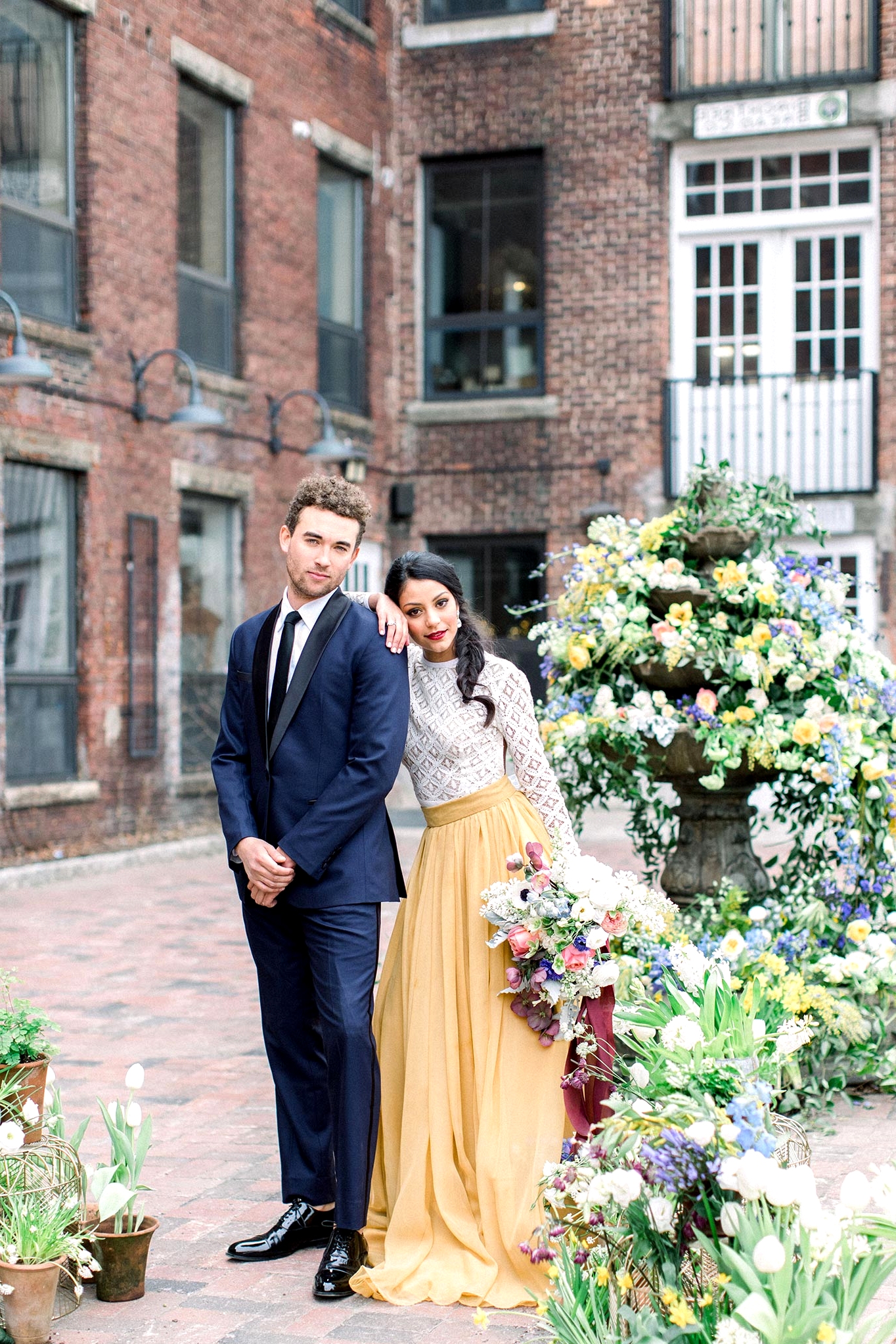 courtyard wedding ceremony with potted plant aisle markers, a floral fountain backdrop, a groom in a navy suit and bride in a two piece wedding dress with a mustard skirt and lace top