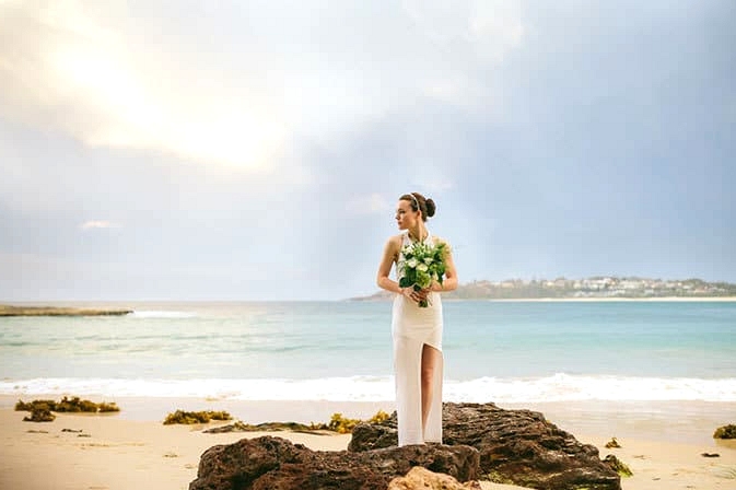 Bride in modern asymmetrical wedding dress standing on beach with bouquet