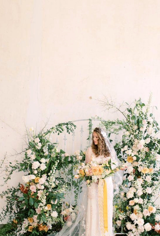 lush floral gate wedding backdrop with bride in a vintage-inspired wedding dress holding a yellow and white spring bridal bouquet