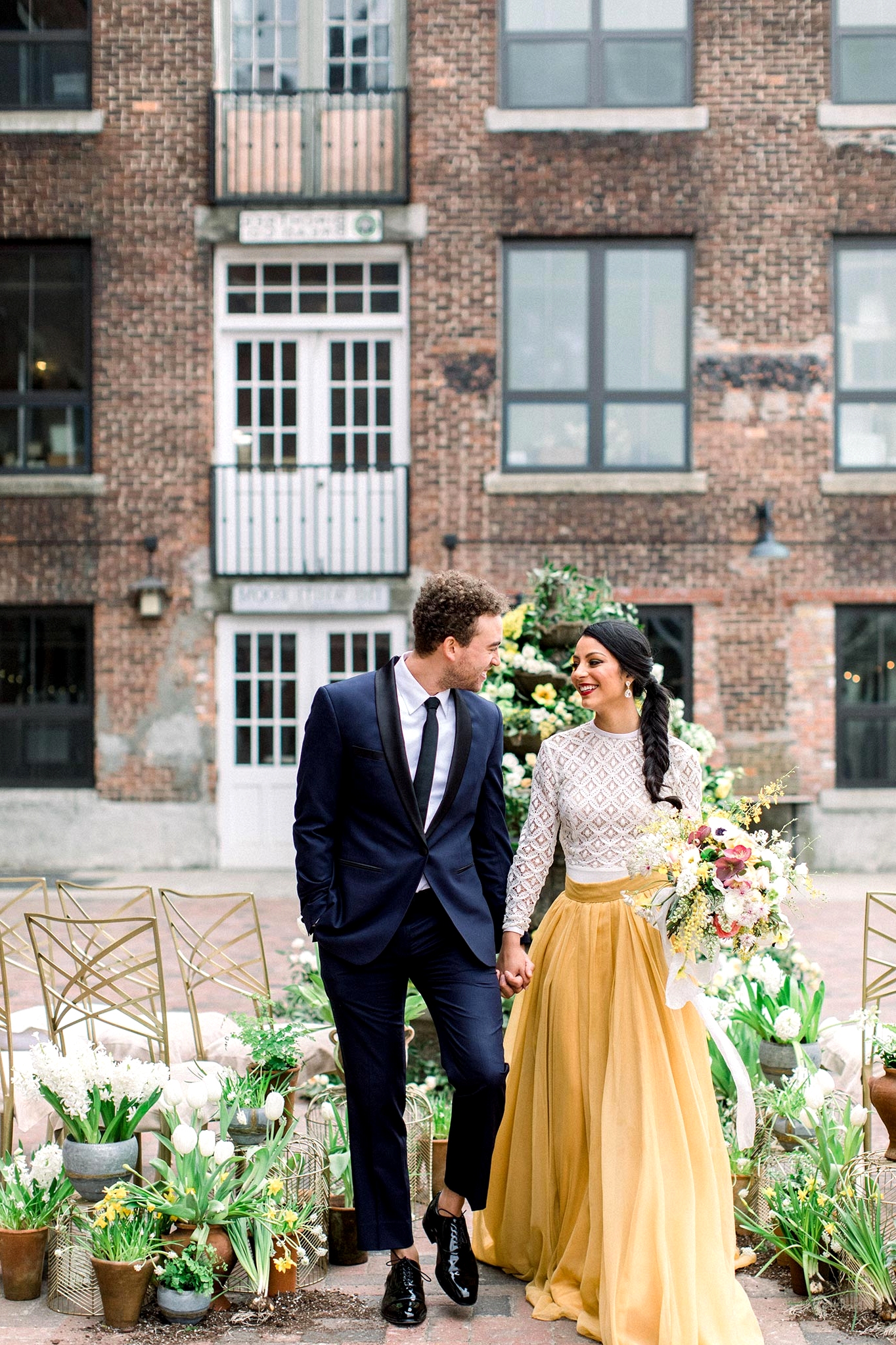 courtyard wedding ceremony with potted plant aisle markers, a floral fountain backdrop, a groom in a navy suit and bride in a two piece wedding dress with a mustard skirt and lace top